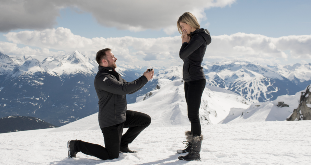 A person kneels in the snow while proposing to another person standing in front of them. Both are dressed in winter clothing, surrounded by a snowy mountainous landscape with clear skies and scattered clouds. The standing person appears surprised and emotional.