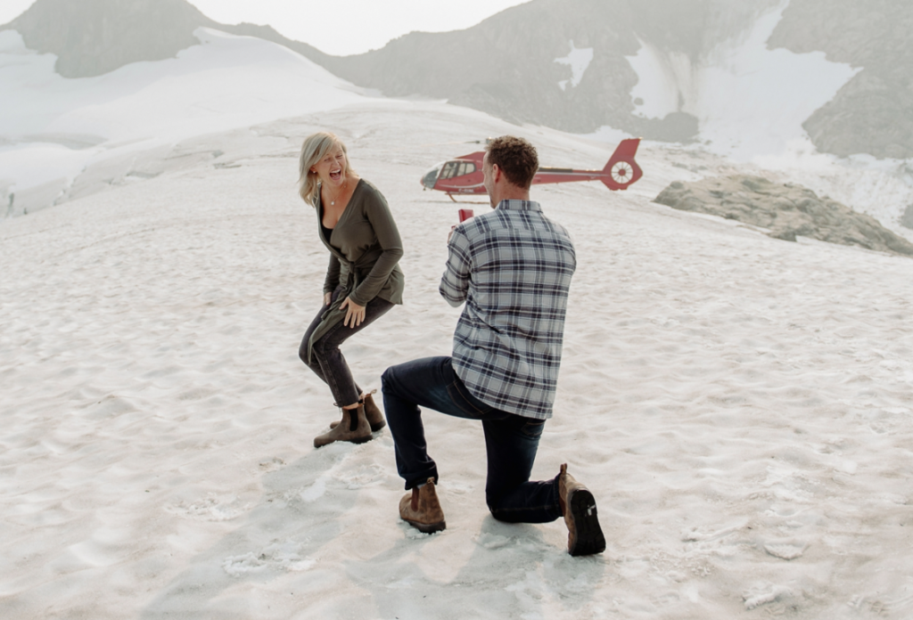 A man kneels and proposes to a woman on a snowy mountaintop. The woman smiles in surprise. A red helicopter rests in the background on the snow. Both are warmly dressed in boots and jackets. Snowy peaks and rocks surround the scene, creating a breathtaking backdrop.