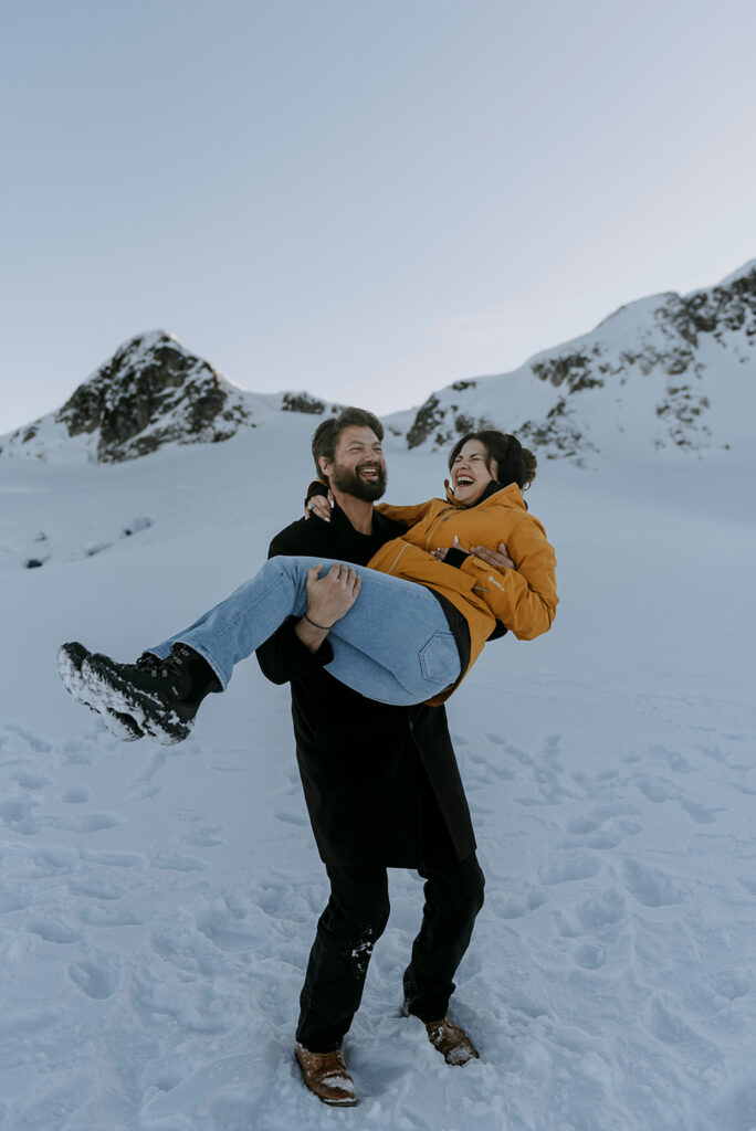 A man carries his fiancé, surrounded by snow.
