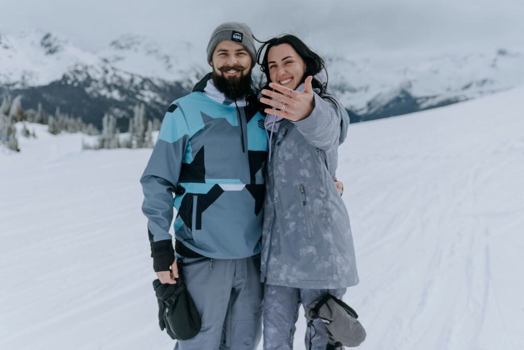 Woman holds up new engagement ring on mountain in snowboard clothing