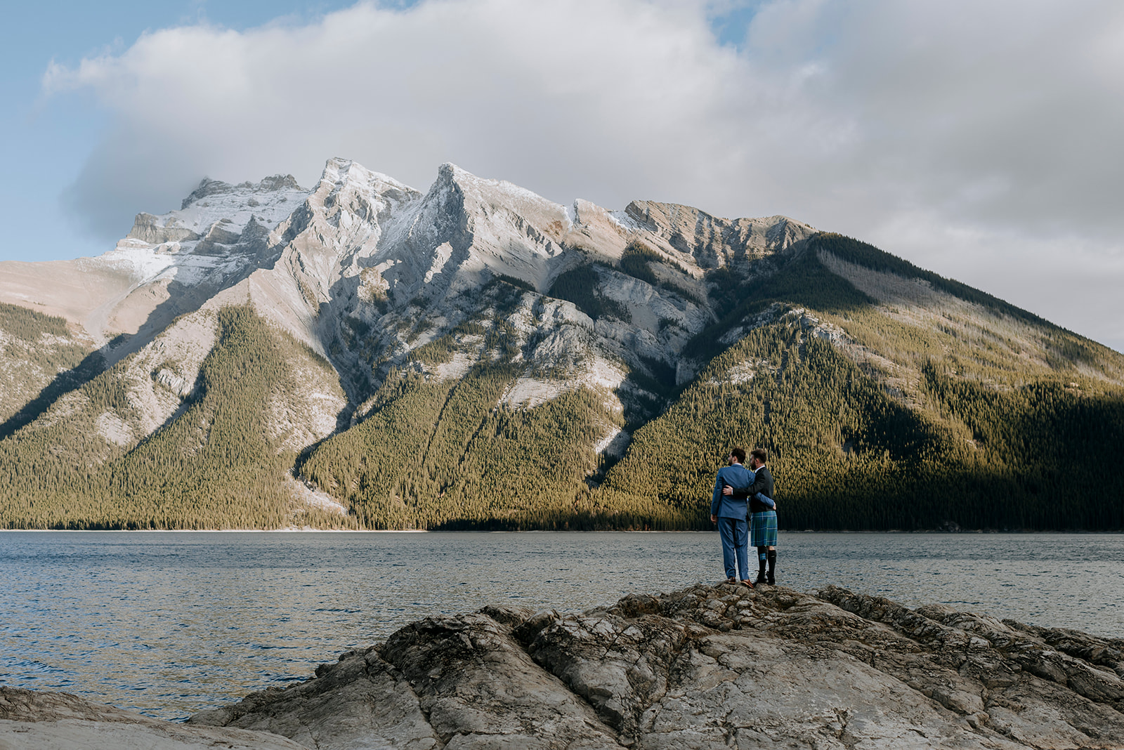 Two grooms dressed in a blue suit and kilt stand in front of Minnewanka Lake in Banff, looking out to the mountain view.
