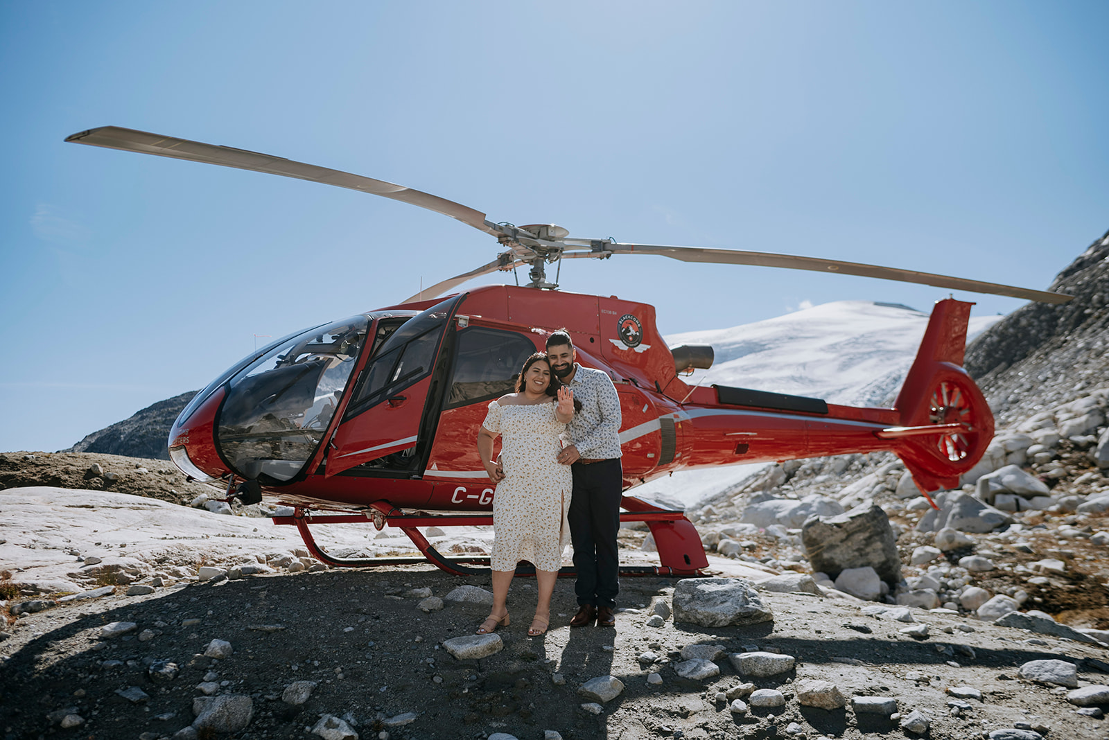 A couple in smart casual stand outside a red helicopter on a mountain on a sunny day. There is snow and glacier in the background.