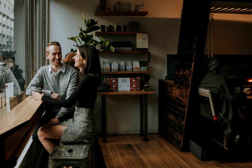 A man and a woman sit at a window-side counter in a cozy Vancouver cafe. They are smiling and engaged in conversation. Shelves with various items and potted plants create a charming backdrop, while another person is seen in the back.