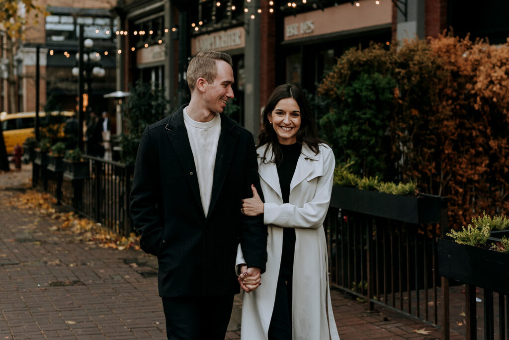 A couple strolls down a brick-paved sidewalk in Vancouver's Gastown, lined with greenery and string lights. The woman, wearing a light-colored coat, smiles while holding the arm of the man, dressed in a dark coat. They seem happy and engaged with each other on a cool, overcast day.