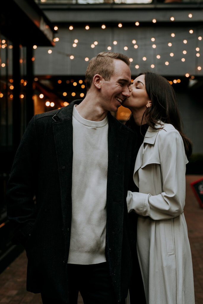 A couple stands close to each other under string lights at night, with the woman kissing the man's cheek. Both are wearing coats; the man in a black coat over a white sweater, and the woman in a light-colored coat. They appear happy and affectionate, capturing their Gastown engagement in Vancouver.