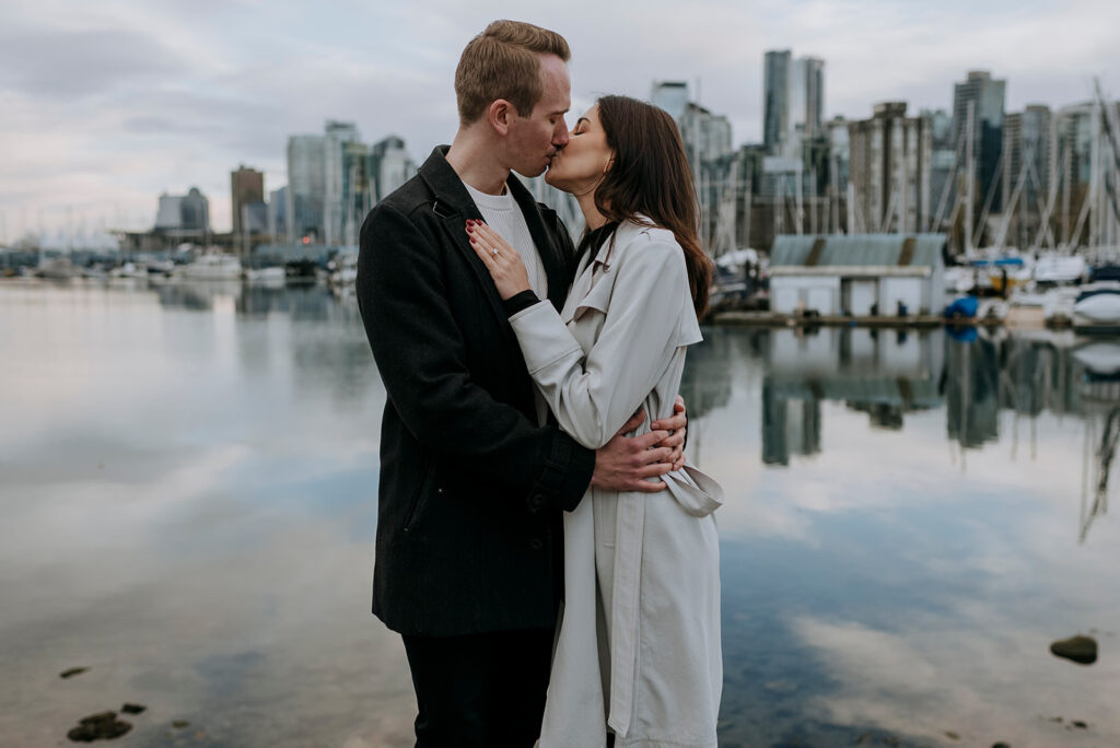 A couple kisses while standing by a marina with the Vancouver skyline in the background. The man is wearing a dark coat, and the woman is wearing a light trench coat. The water reflects the boats and buildings, creating a serene scene perfect for an engagement moment.