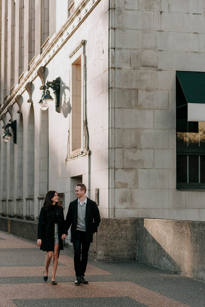 A couple walks hand-in-hand on a city sidewalk in Gastown, Vancouver, next to an ornate building with large windows and decorative wall sconces. Both are dressed in dark coats, and the late afternoon light casts a warm glow on the scene, perfect for an engagement shoot.