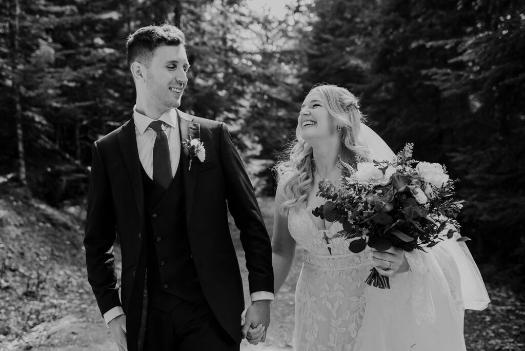 A bride and groom hold hands walking through a forest trail in Whistler. The bride is smiling and grinning at the groom and holds a bouquet of flowers. The image is in black and white.