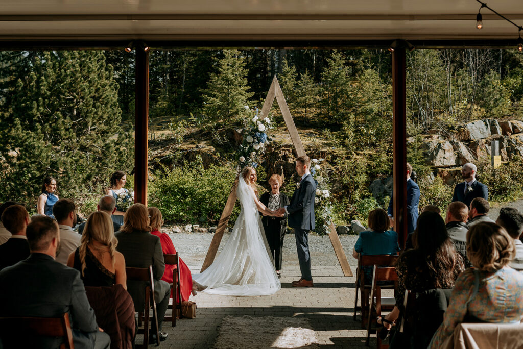 A bride and groom hold hands in their wedding ceremony the Squamish Lil'wat Cultural Centre (SLCC) in Whistler. An officiant wearing black and white stands behind them under an A-frame structure with white and blue flowers. Guests are seated, watching on.