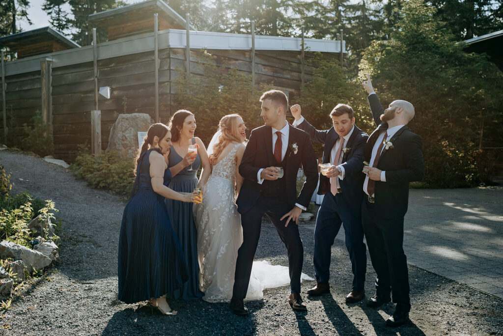 A wedding party dance with each other holding drinks. They're in the sun with a timber structure behind and trees. Two bridesmaids wear blue dresses and two groomsmen wear suits. The bride wears a lace dress and veil and groom wears a suit and red tie.