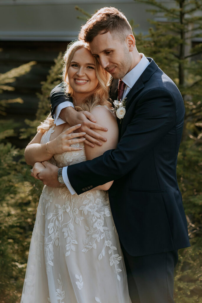 A bride and groom hugging with trees behind. The bride is wearing a lace dress and looking and smiling at the camera. The groom hugs the bride from behind with his right arm over her shoulder and chest.