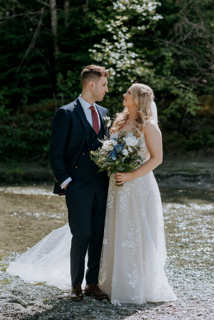 A bride and groom look at each other, surrounded by a river and trees. It is a sunny day. The bride holds a bouquet of white and blue flowers and wears a veil. The groom wears a dark blue suit and red tie.